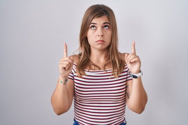 Young hispanic woman standing over isolated background pointing up looking sad and upset, indicating direction with fingers, unhappy and depressed. 