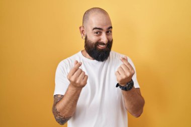 Sakallı ve dövmeli genç bir İspanyol. Sarı arka planda duruyor. Elleriyle para hareketi yapıyor. 