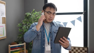 Young chinese man preschool teacher using touchpad talking on smartphone at kindergarten
