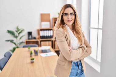 Genç, sarışın, iş kadını, gülümseyen, kendine güvenen, kollarını kavuşturup ofiste el kol kola duran.