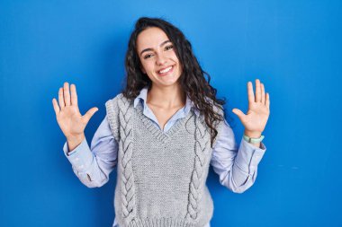 Genç esmer kadın mavi arka planda duruyor ve 10 numaralı parmaklarıyla işaret ederken kendinden emin ve mutlu gülümsüyor.. 