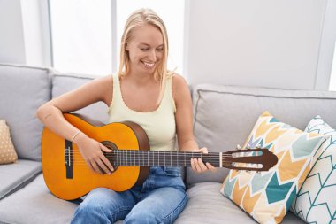 Genç sarışın kadın evdeki kanepede oturmuş klasik gitar çalıyor.