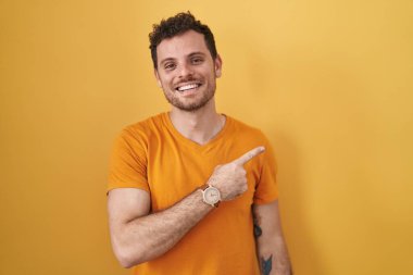 Young hispanic man standing over yellow background cheerful with a smile on face pointing with hand and finger up to the side with happy and natural expression 