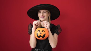 Young blonde woman wearing witch costume holding pumpkin basket over isolated red background