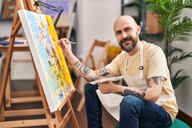 Sanat stüdyosunda kendine güvenen genç kel sanatçı gülümsüyor.