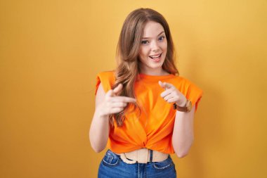 Sarı arka planda dikilen beyaz kadın mutlu ve komik yüzlü parmaklarını kameraya doğrultuyor. iyi enerji ve titreşimler. 