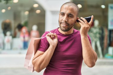 Young latin man listening voice message by smartphone holding shopping bag at street
