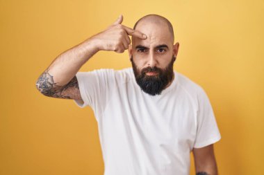 Sarı arka planda dikilen sakallı ve dövmeli genç İspanyol adam alnındaki sivilceden mutsuz olduğunu gösteriyor. Siyah noktalı çirkin bir enfeksiyon. sivilce ve deri problemi 