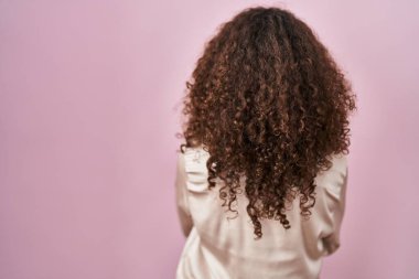 Pembe arka planda duran kıvırcık saçlı İspanyol kadın kollarını kavuşturup arkaya bakıyor. 