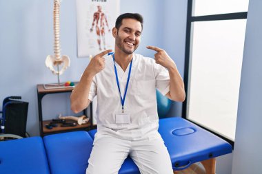 Sakallı, sakallı, acılı bir klinikte çalışan neşeli gülümseyen ve parmaklarıyla ve ağzıyla işaret eden genç bir İspanyol. diş sağlığı kavramı. 