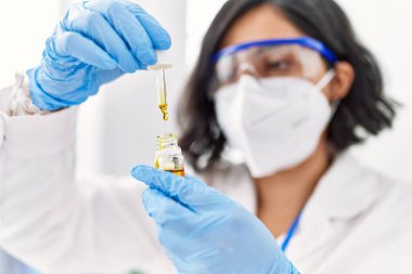 Young latin woman wearing scientist uniform and medical mask using pipette at laboratory