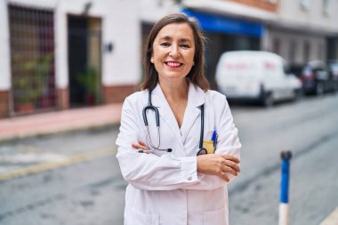 Middle age woman wearing doctor uniform standing with arms crossed gesture at street