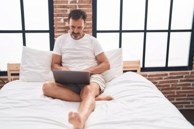 Middle age man using laptop sitting on bed at bedroom