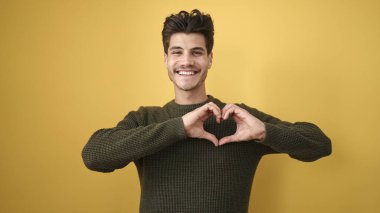 Young hispanic man smiling confident doing heart gesture with hands over isolated yellow background