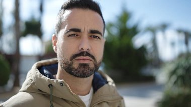 Young hispanic man standing with serious expression at park