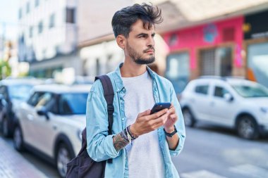 Young hispanic man student using smartphone with relaxed expression at street