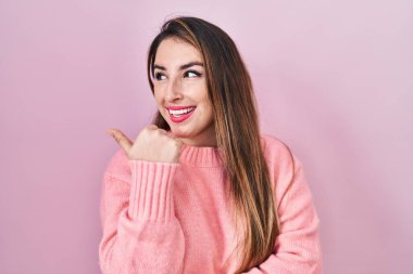 Young hispanic woman standing over pink background smiling with happy face looking and pointing to the side with thumb up. 