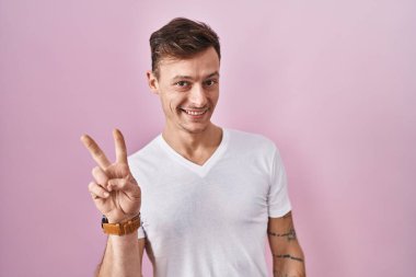 Pembe arka planda duran beyaz bir adam kameraya gülümseyen bir gülümsemeyle parmaklarıyla zafer işareti yapıyor. İki numara.. 