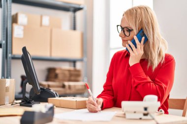 Genç sarışın kadın ecommerce iş işçisi ofisteki belgelerde akıllı telefon yazısıyla konuşuyor.