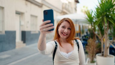 Genç kızıl saçlı kadın gülümsüyor. Kendine güveni tam. Sokakta akıllı telefonun yanında selfie çekiyor.