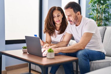 Orta yaşlı erkek ve kadın çift evdeki koltukta dizüstü bilgisayar ve kredi kartı kullanıyorlar.