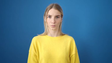 İzole edilmiş mavi arka planda ciddi bir ifadeyle duran genç sarışın kadın.