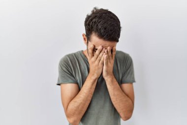 Beyaz arka plan üzerine gündelik tişört giyen sakallı genç İspanyol bir adam. Ağlarken suratını ellerle kapatan üzgün bir ifade. depresyon kavramı. 