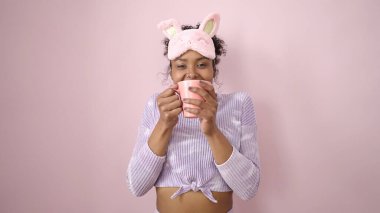 African american woman wearing sleep mask drinking cup of coffee over isolated pink background