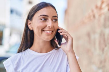 Genç, güzel, İspanyol bir kadın gülümsüyor ve caddede akıllı telefondan konuşuyor.