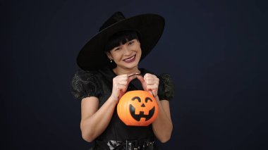 Young chinese woman wearing witch costume holding halloween pumpkin basket over isolated black background