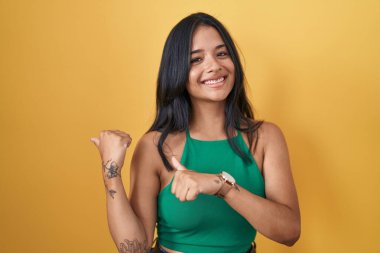 Sarı arka planda duran esmer kadın, arka tarafı işaret ediyor, baş parmakları havada, kendinden emin gülümsüyor. 