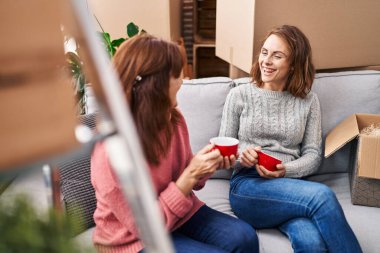 Two women mother and daughter drinking coffee sitting on sofa at new home