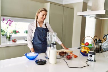 Genç sarışın kadın gülümsüyor. Mutfakta kendine güvenen smoothie pişiriyor.
