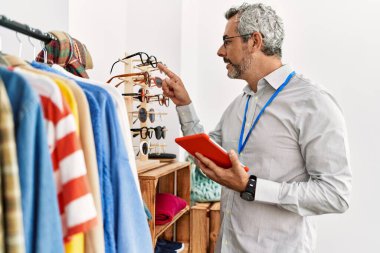 Orta yaşlı, gri saçlı bir dükkan asistanı giyim mağazasında dokunmatik ped kullanıyor.