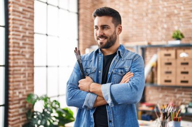 Genç İspanyol adam gülümsüyor. Kollarını kavuşturup sanat stüdyosunda duruyor.