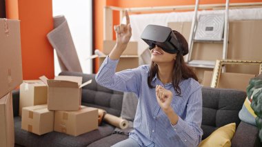 Sanal gerçeklik gözlüğü takan genç, güzel İspanyol kadın yeni evindeki kanepede oturuyor.