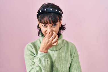 Young beautiful woman standing over pink background smelling something stinky and disgusting, intolerable smell, holding breath with fingers on nose. bad smell 