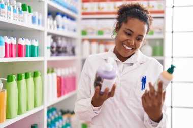 African american woman pharmacist smiling confident holding gel bottles at pharmacy