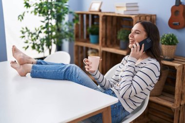 Akıllı telefondan konuşan, evde kahve içen genç bir kadın.