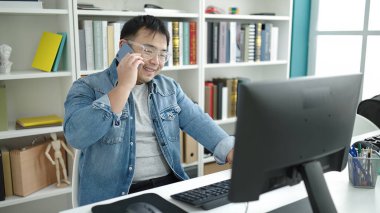 Young chinese man student using computer talking on smartphone at library university
