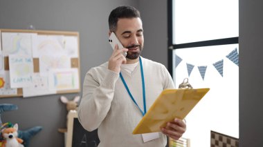 Young hispanic man preschool teacher reading document talking on smartphone at kindergarten
