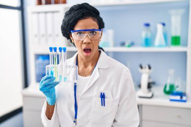 Middle age hispanic woman working at scientist laboratory scared and amazed with open mouth for surprise, disbelief face 