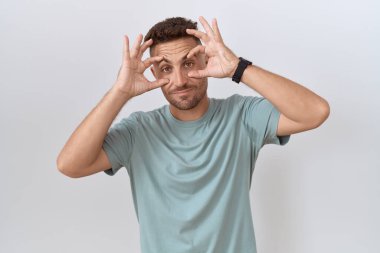 Hispanic man with beard standing over white background trying to open eyes with fingers, sleepy and tired for morning fatigue 