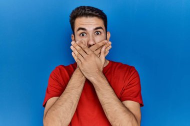 Mavi arka planda kırmızı tişört giyen, sakallı, ağzı yanlışlıkla ellerle kapatan İspanyol genç bir adam. gizli kavram. 