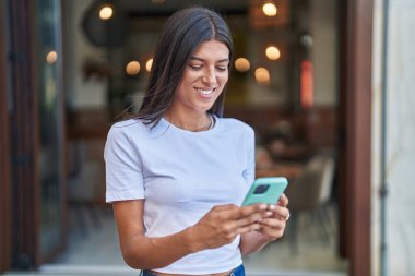 Genç, güzel, İspanyol bir kadın sokakta akıllı telefon kullanarak gülümsüyor.