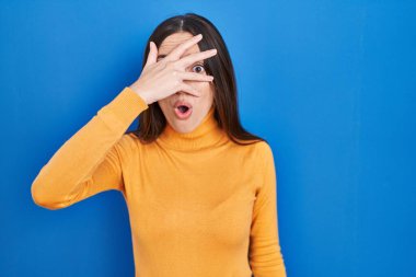 Genç esmer kadın mavi arka planda dikiliyor. Şoka girip yüzünü ve gözlerini kapatıyor. Utanmış bir ifadeyle parmaklarından bakıyor.. 
