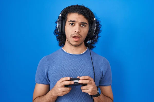 ヒスパニック系の男とともに巻き毛再生ビデオゲームホールディングコントローラーで衝撃的な顔 懐疑的で皮肉 驚きますオープン口 — ストック写真