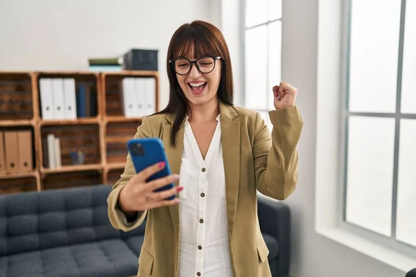 若いブルネットの女性は誇りを持って叫んでスマートフォンでオフィスで働いています 勝利と成功を祝います非常に腕を上げ興奮 — ストック写真