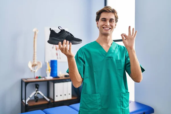 Young Man Working Physiotherapy Clinic Holding Shoe Doing Sign Fingers — Stock Photo, Image