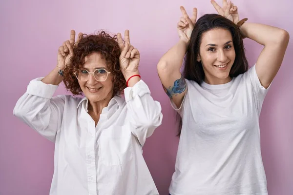 ヒスパニック系の母親と娘身に着けていますカジュアルホワイトTシャツ上のピンクの背景Posing面白いとクレイジーとともに指で頭にウサギの耳 笑顔陽気に — ストック写真